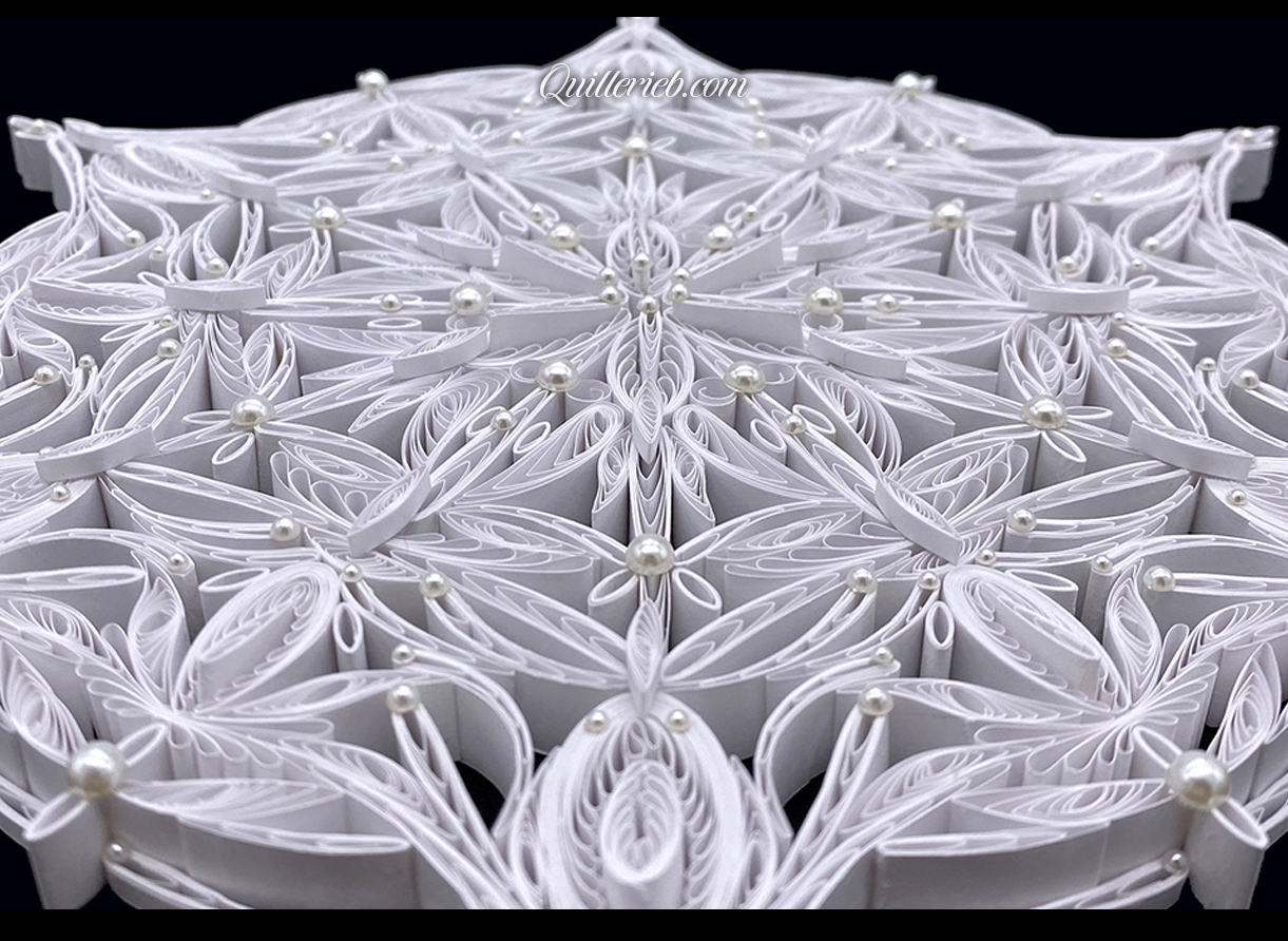 Circular 6-pointed quilling in white, snowflake design close-up slanted backwards view.