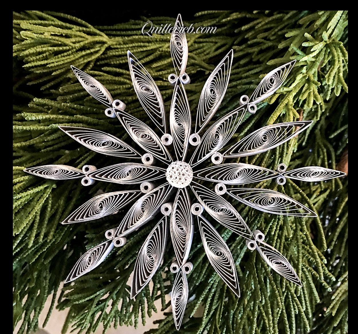 Quilling in white, 3rd snowflake design full view against pine wreath backdrop.