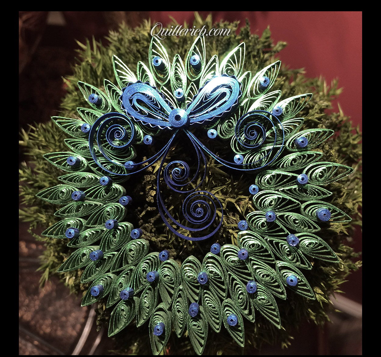  Quilled wreath shape with bow and ribbon, green and blue design full view against pine wreath backdrop.